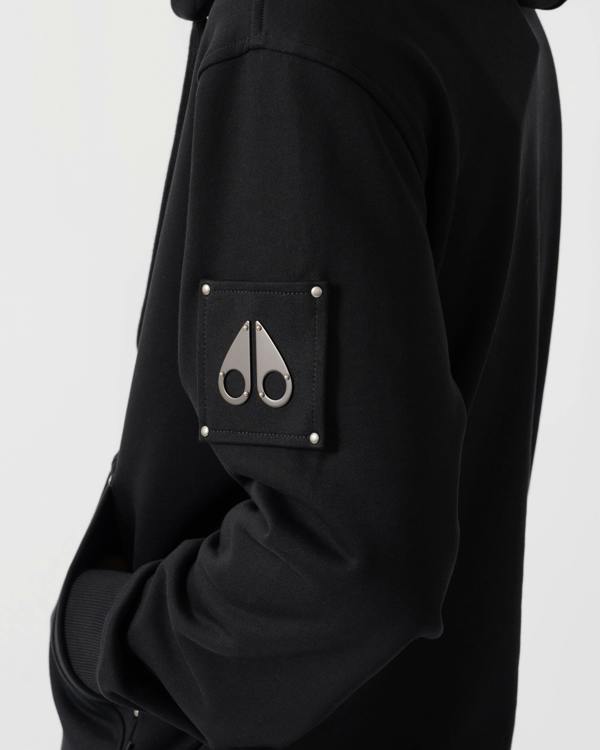 Close-up of a black hooded jacket featuring a square chest pocket with silver rivets and a silver metal scissors logo on the front.