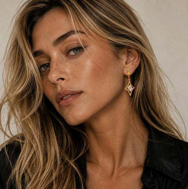 A close-up of a woman with blonde hair wearing a gold dangle earring with a clear rhinestone, paired with a black satin shirt against a neutral beige background.