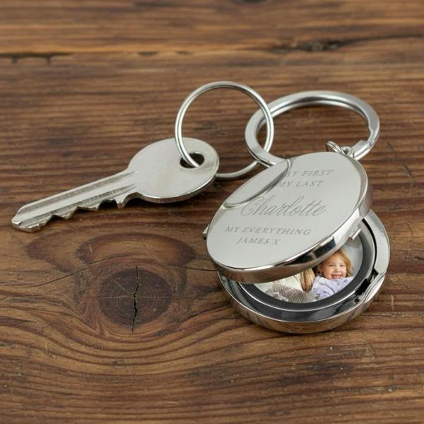A silver, circular personalised photo keyring lying on a wooden surface next to a metal house key. The keyring is open, revealing a photograph of a smiling young child inside.