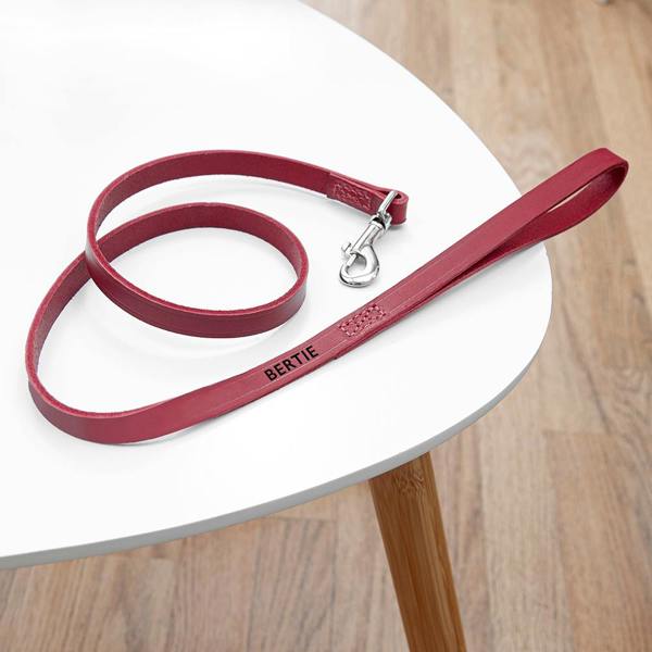A personalised classic red leather dog lead stamped with the name BERTIE in black foil, coiled on a white wooden table.