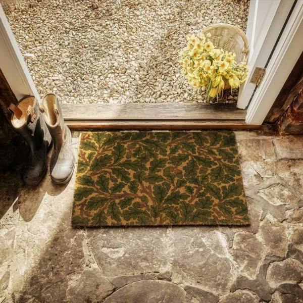 Doormat with leaf pattern on stone floor, shoes, and basket of flowers.