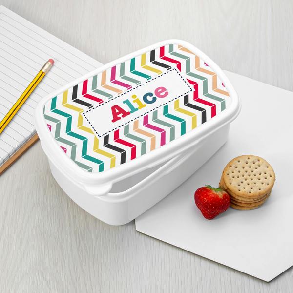 A white plastic personalised girl's lunch box printed with vibrant geometric chevron zigzags and the name Alice, styled next to a notebook, pencil, fresh strawberry, and crackers.