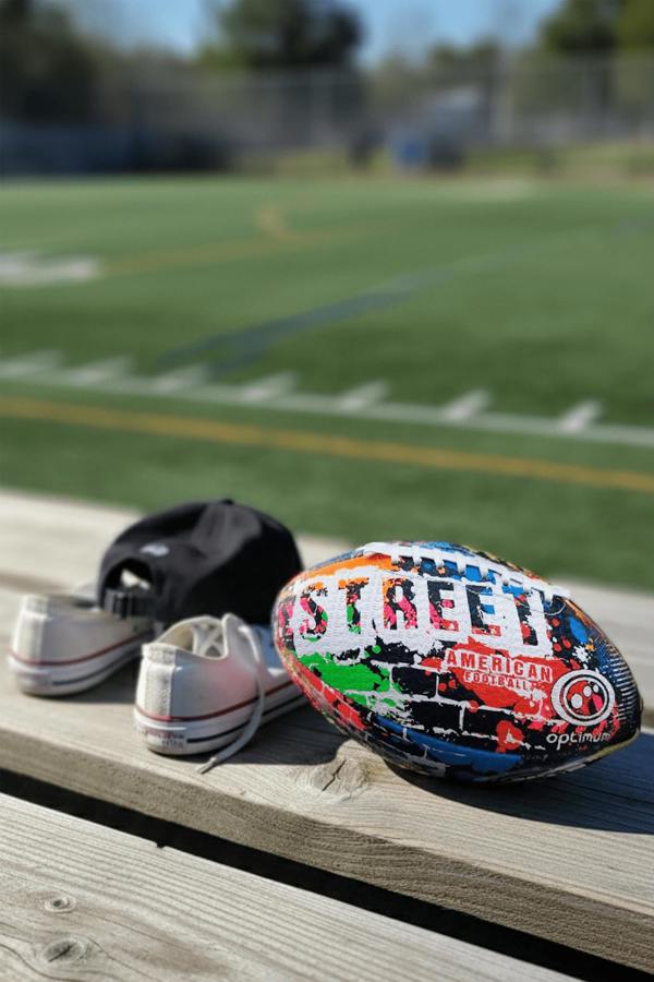 Multicolour Street American football with white laces on wooden bench, cap, trainers and sports field behind - Optimum