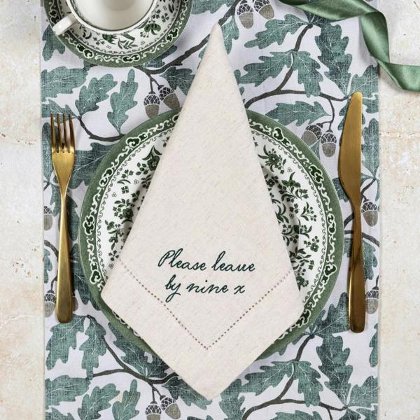 A natural linen napkin folded into a point on a green and white patterned plate, featuring green embroidered script that reads 'Please leave by nine x'.