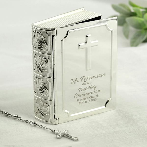 A silver-finish bible trinket box by Abode Home & Gifts, shown standing slightly open on a white linen surface. The cover is engraved with "Ida Rosemarie On Your First Holy Communion St Peter's Church 23rd July 2023", next to a silver rosary chain.