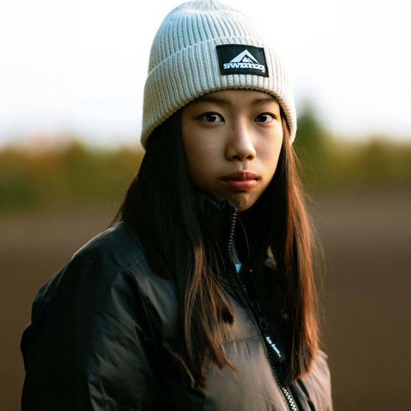 Young woman wearing a simple beanie hat knitted in a chunky single rib in an oatmeal shade. There is a SWIMZI badge sewn to the front of the deep turn back headband.