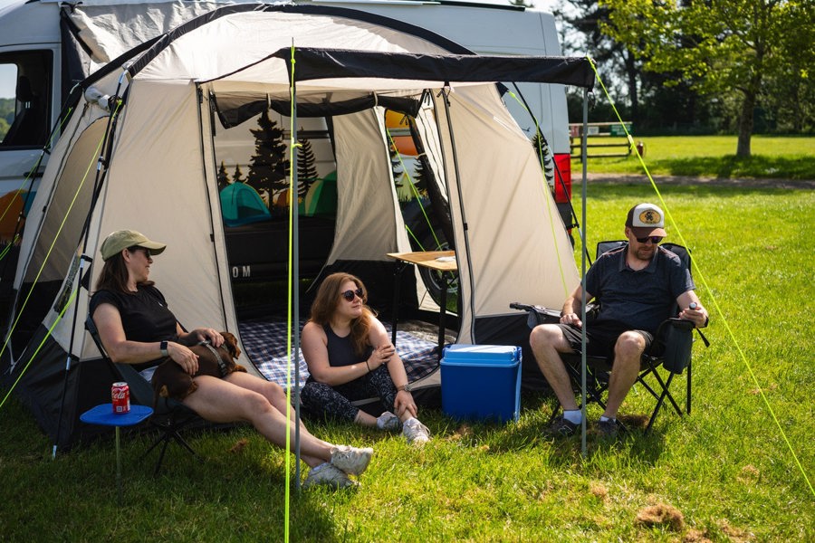 A group of people relaxing outside of the Cubo XL Motorhome Awning - Fibreglass Poles that is attached to a Volkswagen Crafter.