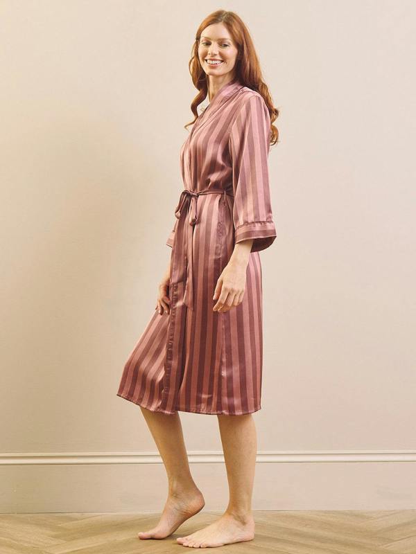 A woman modeling a satin-like robe with vertical stripes in dusty rose and dark brown, standing barefoot against a plain beige wall with a light wood floor.
