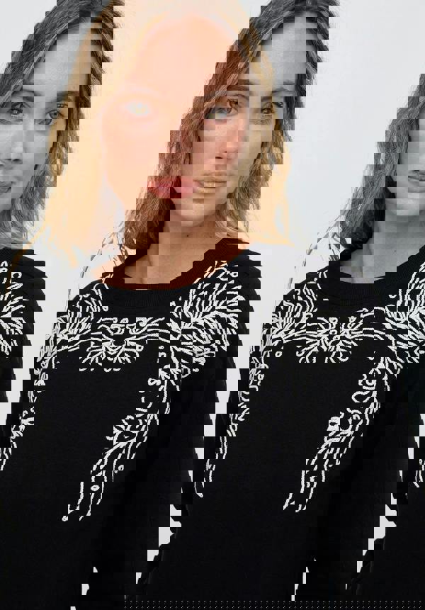 Woman wearing a black sweater with white floral embroidery on a plain background