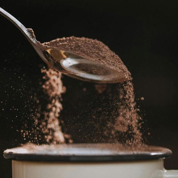 Fine artisan hot chocolate powder cascading from a silver metal spoon into an enamel mug against a dark backdrop.