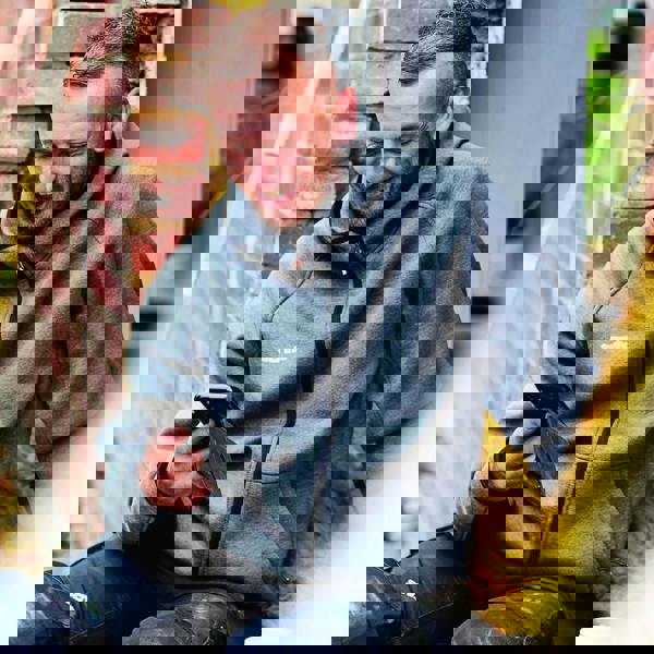 Scruffs Trade Air Mens Sweat Shirt Hooded Full Zip Hoodie, a man wearing a grey hooded zip-up sweatshirt styled with black accents, sitting outdoors near a brick wall, looking at his phone and smiling.