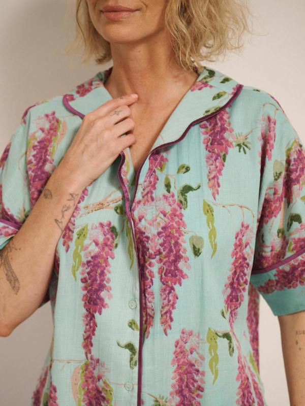 Woman wearing duck egg blue cotton gauze oversized pyjamas with pink wisteria floral print, short-sleeve button-through top and wide-leg trousers, standing barefoot indoors.