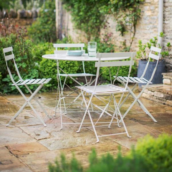 A French-inspired chalk white metal bistro set with a round table and four folding chairs on a wet stone patio next to an old stone wall.