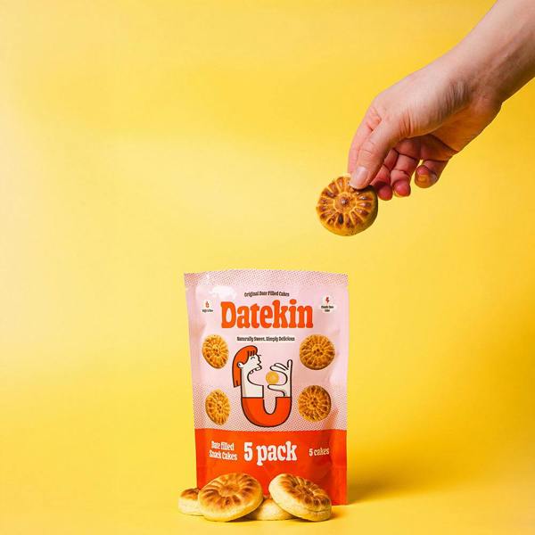 Hand holding a Datekin date snack cake above its packaging on a yellow background