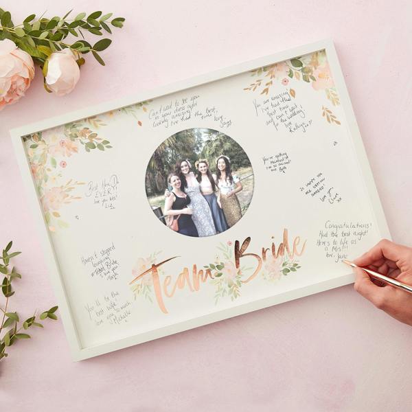 Top-down view of a white signature guest book frame with signed messages and a rose gold foiled Team Bride mount as a hand writes on it.