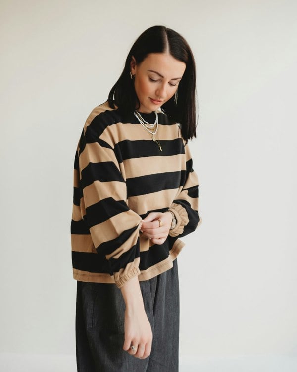 A woman with shoulder-length dark hair stands against a plain background, wearing the Ivy Striped Sweatshirt in beige and black made of organic cotton and dark pants. She appears to adjust her balloon sleeve while accessorizing with layered necklaces and hoop earrings.