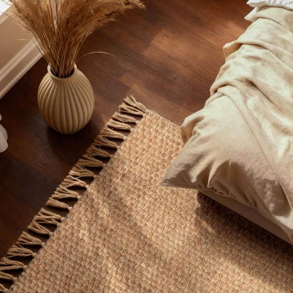 Beige pillow on a textured rug next to a vase with dried plants on a wooden floor.