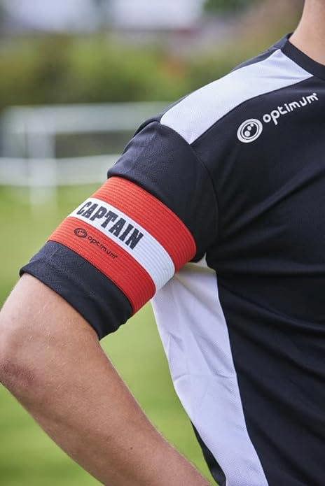 Red Optimum captain's armband worn on a player's arm over black and white kit on a pitch - Optimum