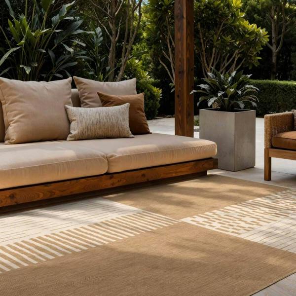 Outdoor seating area with a beige sofa and cushions on a patterned rug, surrounded by plants.