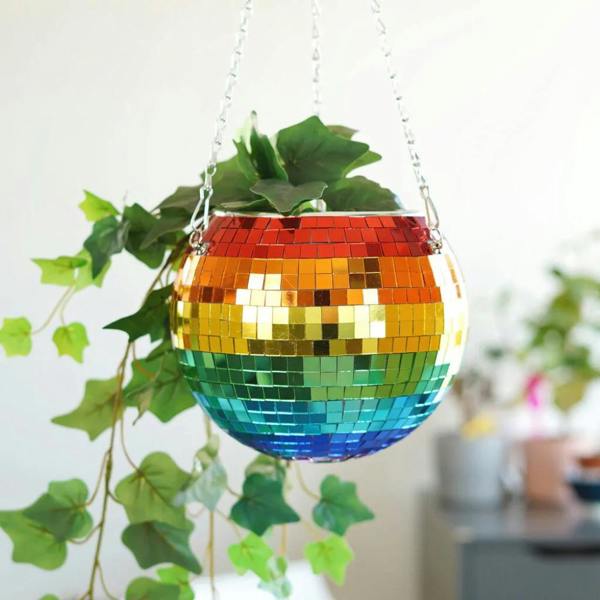 A colourful rainbow mirrored disco ball hanging planter filled with a green trailing ivy plant, suspended by a silver metal chain in front of a white wall.