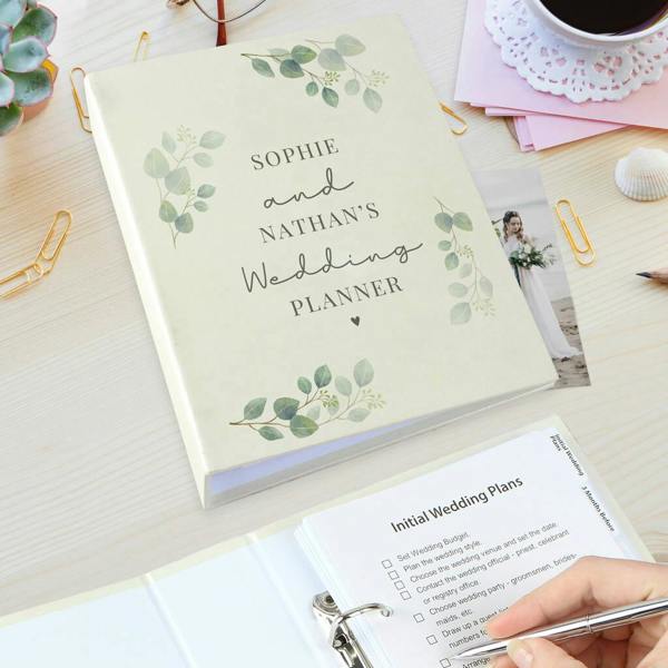 A personalised wedding planner with a cream cover and green eucalyptus leaves reading Sophie and Nathan's Wedding Planner, shown next to an open binder showing an initial wedding plans checklist.