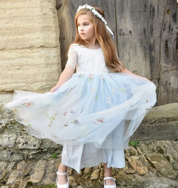 Young girl in a white dress with a light blue floral skirt standing against a stone wall.