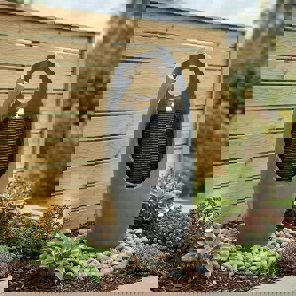 Sleek gray electric water feature with cascading water, illuminated by a soft light, surrounded by smooth pebbles and lush greenery