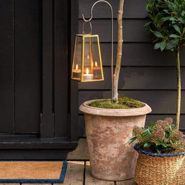 Small raw metal festoon hook styled in a terracotta pot, holding a gold glass lantern with flickering candles against a dark wood-cladded wall.