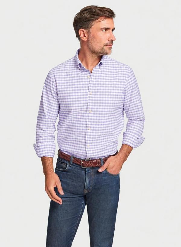 A man wearing a light purple and white gingham long-sleeve button-down shirt, paired with dark blue jeans and a brown belt, standing against a light gray background.