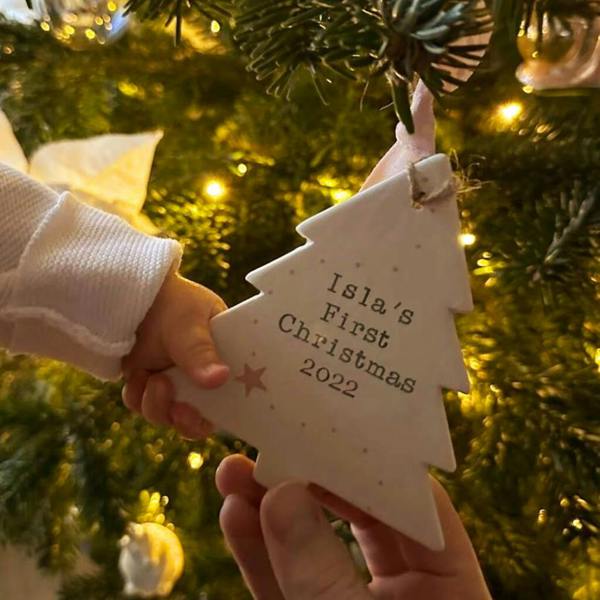 A close-up of a baby's hand touching a white ceramic tree ornament reading 'Isla's First Christmas 2022' hanging on a glowing pine tree.