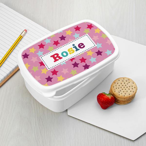 A white plastic personalised girl's lunch box featuring a pink lid with colourful star patterns and the name Rosie, set beside a notebook, pencil, fresh strawberry, and crackers.