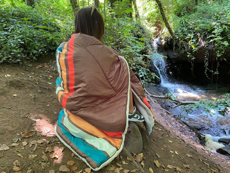 Young lady sat in the outdoors on a log wrapped in a Sherpa lined Vivid Spruce Blanko outdoor Blanket. imageset:Vivid Spruce