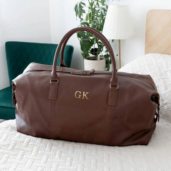 A rich, chocolate-brown vegan leather weekender holdall resting on a white quilted bedspread. The bag is elegantly monogrammed with gold-embossed "GK" initials in the centre. It features sturdy carry handles and a sleek zip closure. A green velvet chair and a bedside lamp are visible in the soft-focus background.