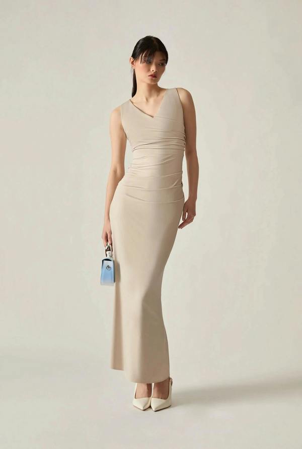 Full‑body shot of a woman wearing a beige dress, holding a small blue bag in her right hand against a soft beige background.