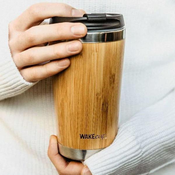 A person wearing a white knit jumper holding a WAKEcup reusable coffee cup made of natural bamboo with a black leak-proof lid and stainless steel trim.