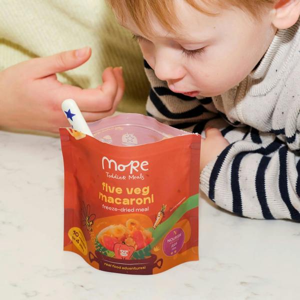 toddler looking at a 'More Toddler Meals Five Veg Macaroni' package on a white surface for dinner.