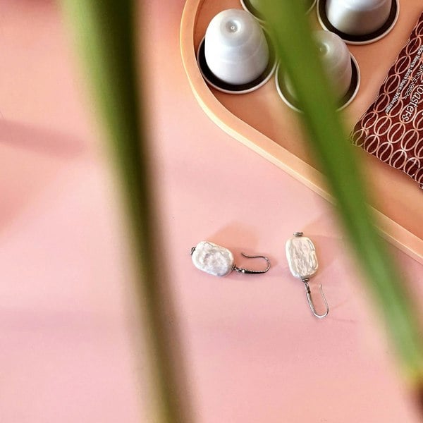 Baroque pearl drop earrings on a pink surface with a plant leaf, coffee pods and a tray.