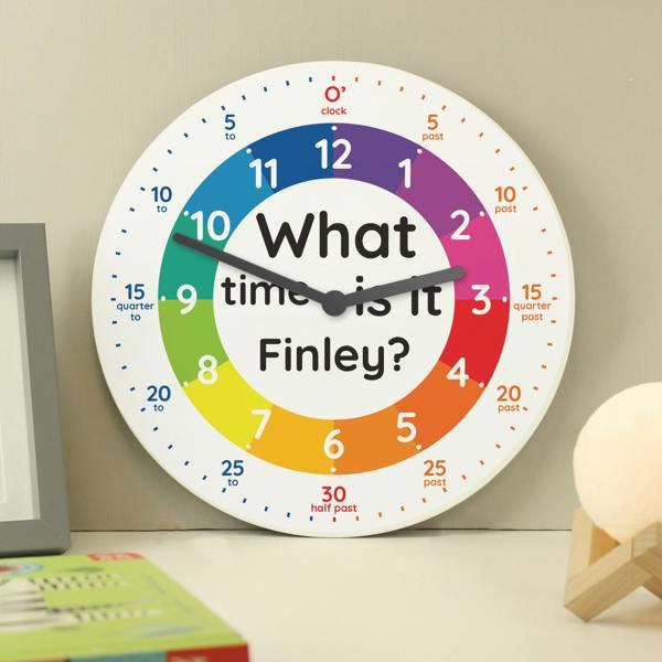 A personalised children's wooden wall clock by Abode Home & Gifts featuring a colourful rainbow time-teaching dial that reads "What time is it Finley?", styled on a desk next to a grey picture frame and a spherical moon lamp.