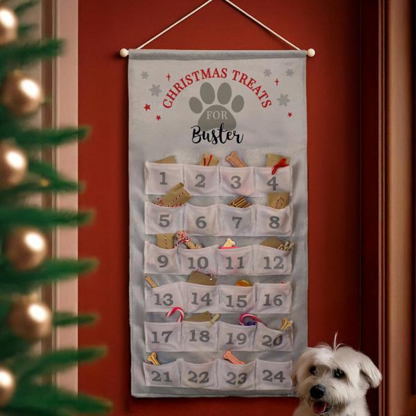 Personalised grey fabric pet advent calendar with 24 numbered pockets holding treats, hanging on a dark red wall next to a white dog.