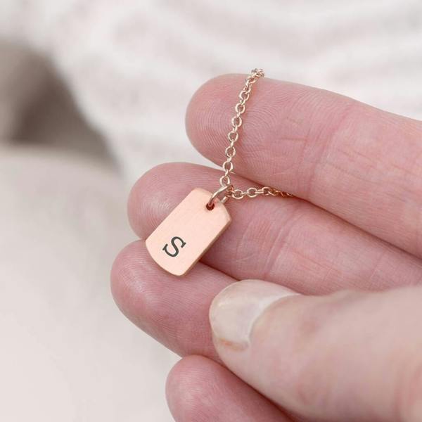 A dainty rose gold-plated rectangular tag pendant necklace engraved with a custom initial letter, displayed on a clean background.