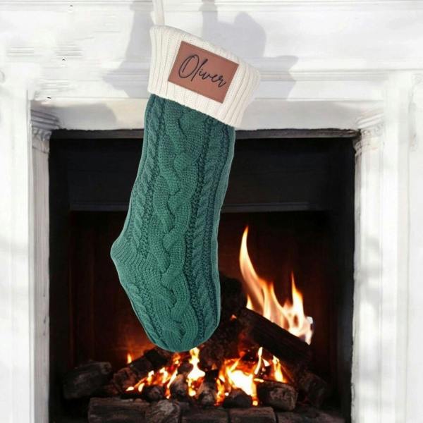 Personalised forest green cable-knit Christmas stocking with an engraved faux leather patch reading Oliver hanging in front of a cosy fireplace.