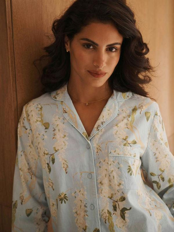 A woman wearing a light blue pyjama shirt with a beige and green floral pattern, leaning against a wooden wall.