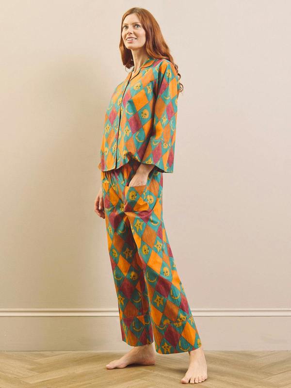 A woman stands barefoot in a room wearing a matching two-piece pyjama set with a colorful geometric ikat pattern in orange, teal, and burgundy.