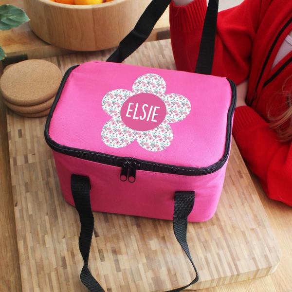 An insulated pink children's lunch bag custom printed with the name Elsie inside a patterned flower illustration, placed on a wooden board next to a person in a red top.