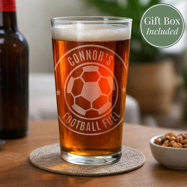 An engraved tulip pint glass by Abode Home & Gifts filled with golden ale and foam, sitting on a wooden table. The glass features a custom football crest reading "BEST DAD EVER" on the top curve and "LOVE HARRY X" on the bottom curve, surrounded by snack bowls.