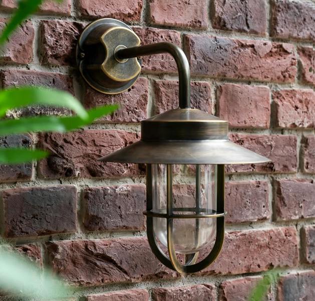 Antique brass outdoor wall light with a storm lamp-inspired cage and clear glass shade, mounted on a red brick wall.