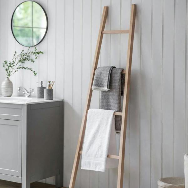 A natural unvarnished beech wood towel ladder leaning against a white shiplap wall in a bathroom, holding white and grey towels.