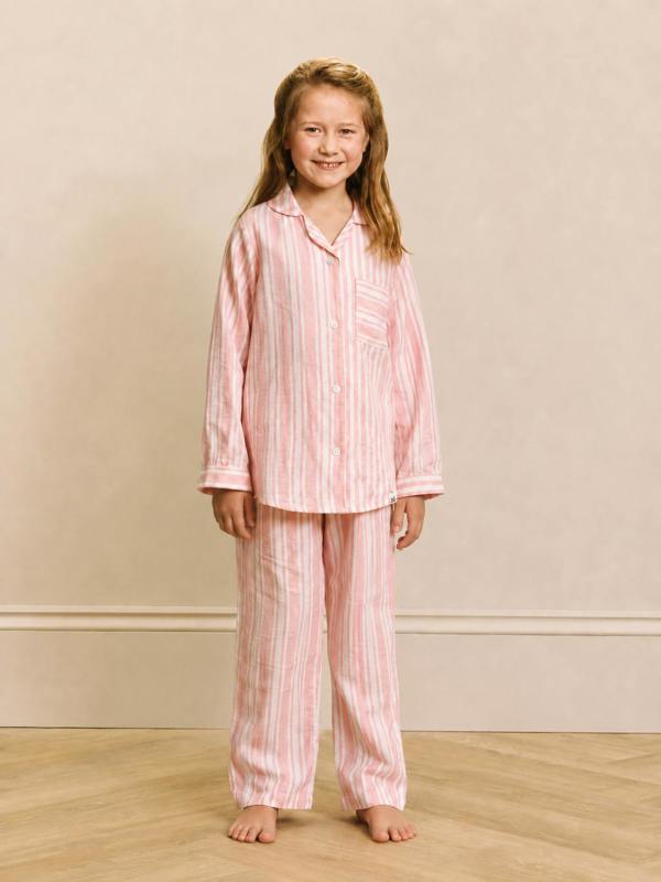 A young girl stands barefoot in a bedroom setting, wearing a two-piece pajama set with vertical pink and white stripes. The set includes a long-sleeve button-up shirt and matching wide-leg pants.