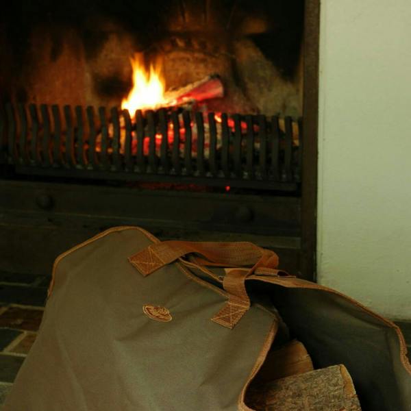 Heavy-duty, dark green canvas log carrier bag with brown handles, filled with chopped firewood and resting on a stone hearth beside a roaring open fireplace with fire tools.