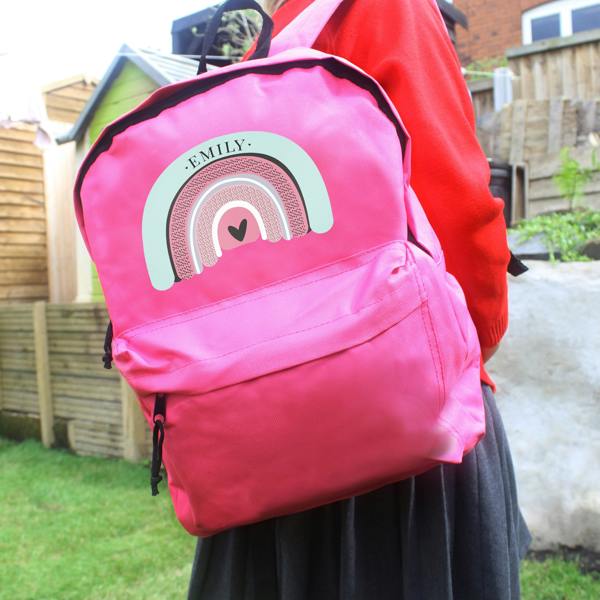 A child wearing a red school uniform sweater and grey skirt holds a personalised pink backpack featuring a pastel rainbow illustration and the name Emily.